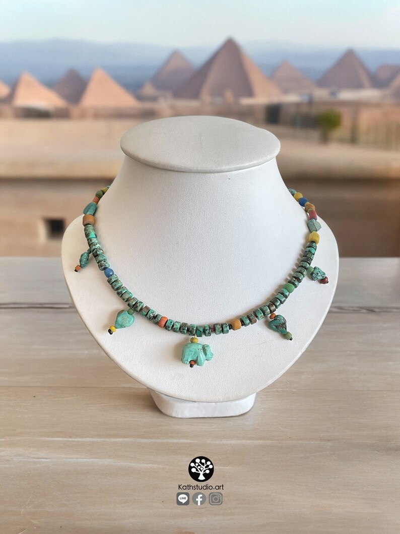 Op de afbeelding: Een turquoise kralenketting met kleine, vierkante kralen en verschillende turquoise bedels in de vorm van vogels en olifanten. De ketting wordt getoond op een witte buste met een vage achtergrond van piramides.
