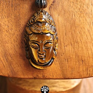 Puede incluir: Un colgante tallado en piedra preciosa ojo de tigre de color marr&oacute;n dorado con forma de rostro sereno, suspendido de un collar de cuentas cil&iacute;ndricas y esf&eacute;ricas de ojo de tigre. El texto Kathstudio.art aparece en la parte inferior.