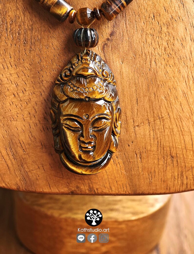 Puede incluir: Un colgante tallado en piedra preciosa ojo de tigre de color marr&oacute;n dorado con forma de rostro sereno, suspendido de un collar de cuentas cil&iacute;ndricas y esf&eacute;ricas de ojo de tigre. El texto Kathstudio.art aparece en la parte inferior.