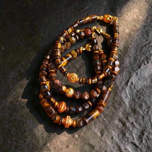 May include: A collection of four bracelets crafted from faceted tiger eye beads in shades of brown and gold. Each bracelet features a central gold-toned metal spacer bead with intricate embossed patterns and a gold-toned metal clasp closure.