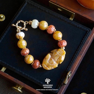 Op de afbeelding: Een armband met ronde kralen in tinten geel, oranje en wit, geaccentueerd met kleine goudkleurige tussenstukjes en een centrale, in de vorm van een blad gesneden gele stenen bedel, voorzien van een goudkleurige knevelsluiting.