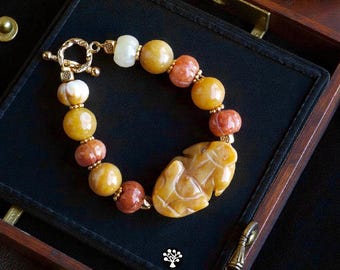 May include: A bracelet featuring round beads in shades of yellow, orange, and white, accented with small gold-toned spacers and a central carved yellow stone charm shaped like a leaf. The piece includes a gold-toned toggle clasp closure.