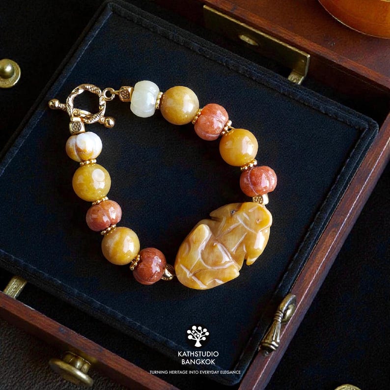 Op de afbeelding: Een armband met ronde kralen in tinten geel, oranje en wit, geaccentueerd met kleine goudkleurige tussenstukjes en een centrale, in de vorm van een blad gesneden gele stenen bedel, voorzien van een goudkleurige knevelsluiting.