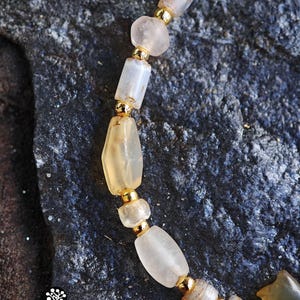May include: A beaded bracelet featuring translucent, irregularly shaped light-colored stones alternating with small, polished gold-tone metal beads. The bracelet includes a carved stone element and displays the text Kathstudio.art with social icons.