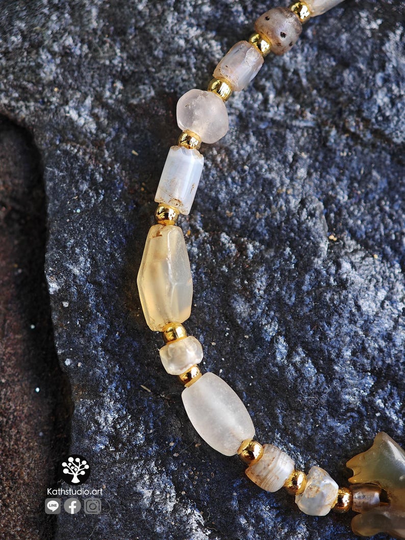 May include: A beaded bracelet featuring translucent, irregularly shaped light-colored stones alternating with small, polished gold-tone metal beads. The bracelet includes a carved stone element and displays the text Kathstudio.art with social icons.