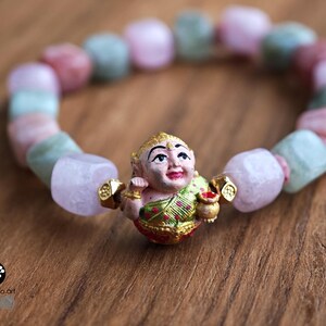 May include: A beaded bracelet featuring a central charm of a painted figure in a green and gold garment holding a small pot, flanked by two gold-toned spacers. The band consists of alternating smooth, irregular-shaped light pink and pale green beads.