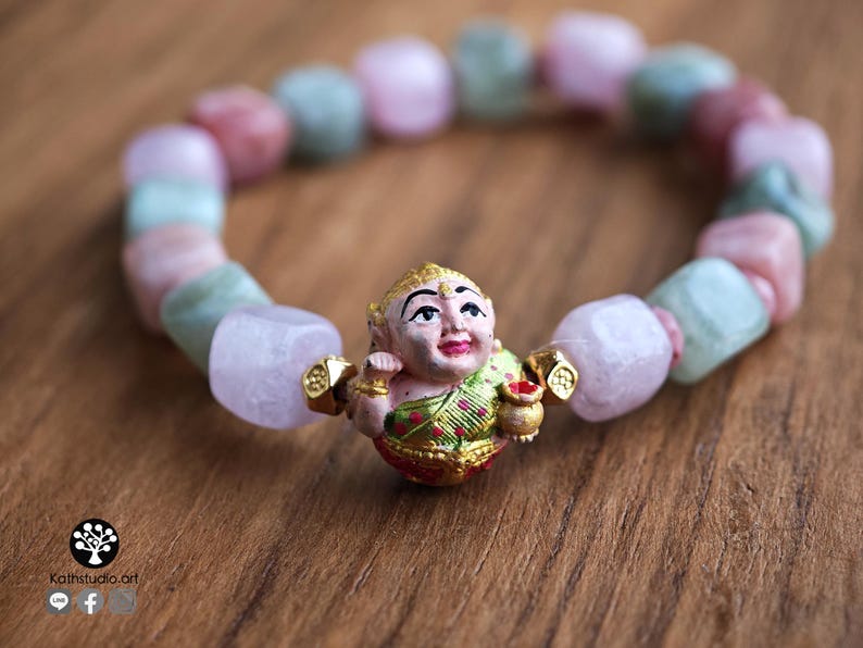 May include: A beaded bracelet featuring a central charm of a painted figure in a green and gold garment holding a small pot, flanked by two gold-toned spacers. The band consists of alternating smooth, irregular-shaped light pink and pale green beads.