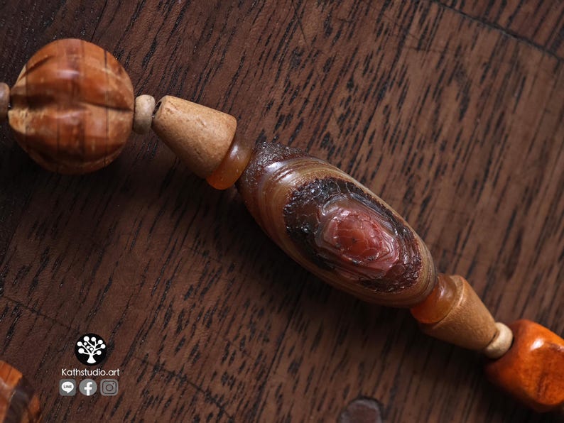 May include: Close-up of a wooden beaded necklace with a carved, textured bead and a central oval bead with a red center. The beads are various shades of brown and orange.