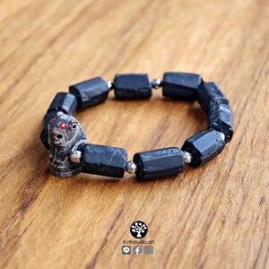 May include: A stretch bracelet featuring cylindrical black tourmaline beads interspersed with small metallic silver spacers. A carved stone animal bead with two small red inlaid eyes is positioned as a focal point on the bracelet.