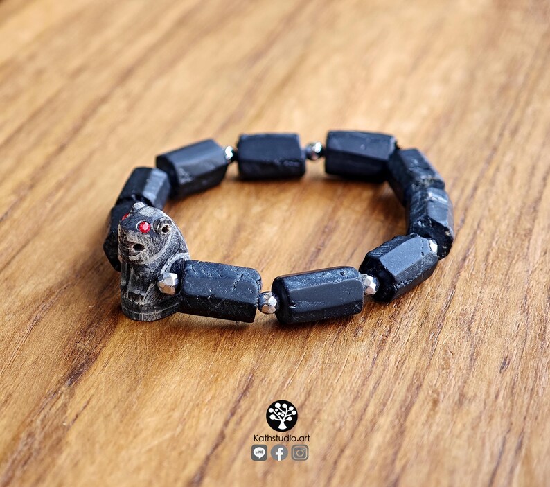 May include: A stretch bracelet featuring cylindrical black tourmaline beads interspersed with small metallic silver spacers. A carved stone animal bead with two small red inlaid eyes is positioned as a focal point on the bracelet.
