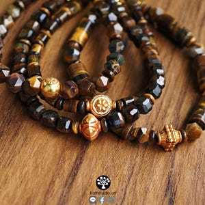 May include: Three beaded bracelets featuring faceted brown and gold tiger eye stones. Each bracelet is accented with a distinct gold-toned metal bead, including one with a floral pattern, one diamond-shaped, and one textured cylindrical bead.