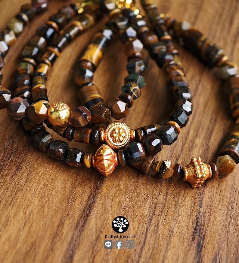 May include: Three beaded bracelets featuring faceted brown and gold tiger eye stones. Each bracelet is accented with a distinct gold-toned metal bead, including one with a floral pattern, one diamond-shaped, and one textured cylindrical bead.