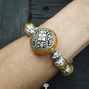 Puede incluir: Una pulsera con cuentas redondas y transl&uacute;cidas de color &aacute;mbar que se alternan con espaciadores de metal plateado ornamentados. La cuenta central es una pieza grande y redonda de color &aacute;mbar con un medall&oacute;n floral de metal plateado.