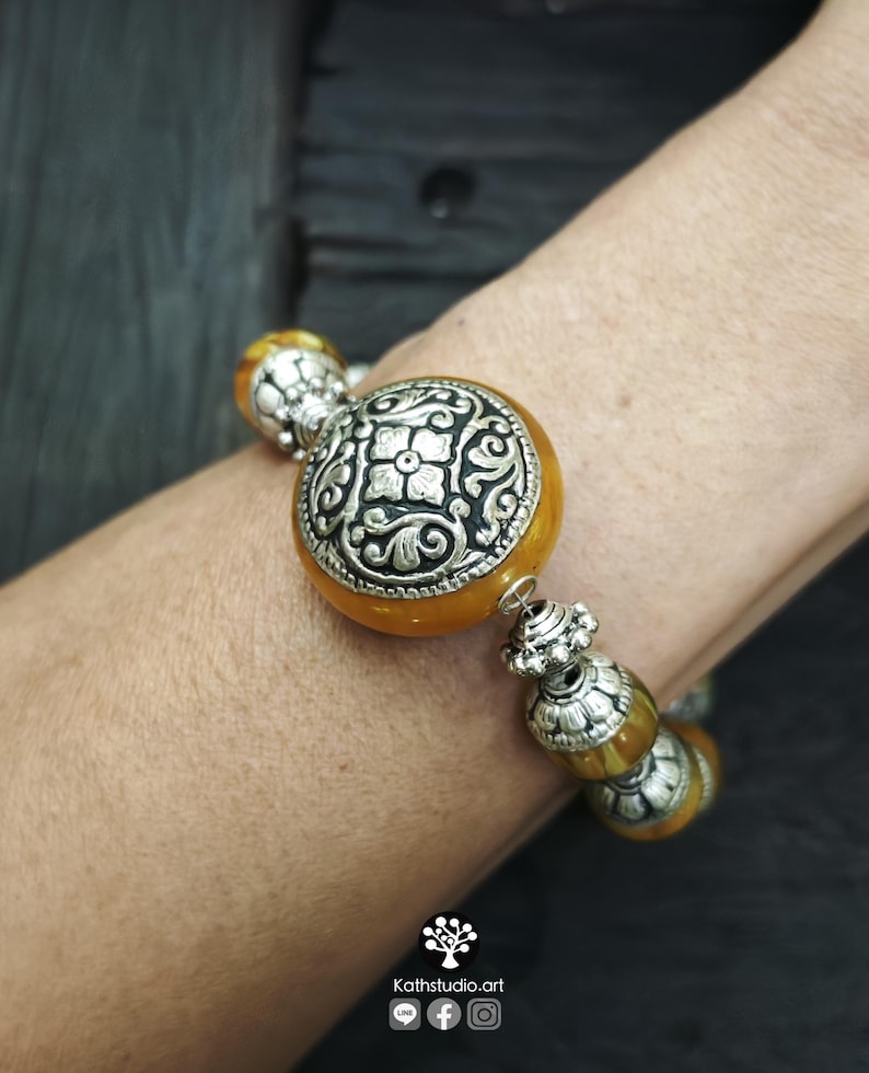 Puede incluir: Una pulsera con cuentas redondas y transl&uacute;cidas de color &aacute;mbar que se alternan con espaciadores de metal plateado ornamentados. La cuenta central es una pieza grande y redonda de color &aacute;mbar con un medall&oacute;n floral de metal plateado.
