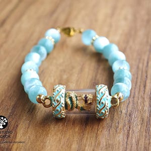 K&ouml;nnte beinhalten: Ein Armband mit hellblauen Katzenaugen-Perlen und einem zentralen, durchsichtigen Glaszylinder, der eine kleine goldfarbene Drachenfigur enth&auml;lt. Der Zylinder wird von zwei t&uuml;rkisfarbenen und goldfarbenen Metall-Abstandhaltern mit Rankenmuster flankiert.