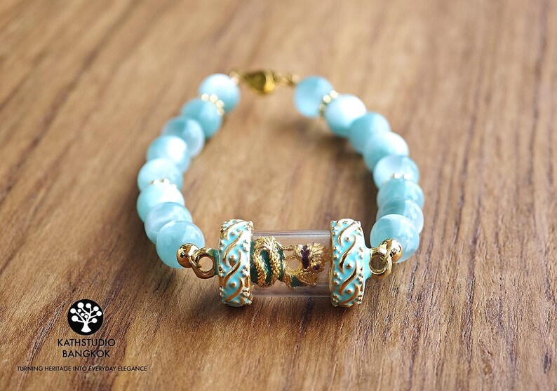 K&ouml;nnte beinhalten: Ein Armband mit hellblauen Katzenaugen-Perlen und einem zentralen, durchsichtigen Glaszylinder, der eine kleine goldfarbene Drachenfigur enth&auml;lt. Der Zylinder wird von zwei t&uuml;rkisfarbenen und goldfarbenen Metall-Abstandhaltern mit Rankenmuster flankiert.