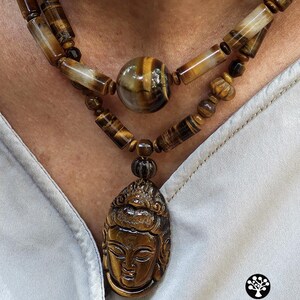 Puede incluir: Un collar de doble hebra con cuentas de ojo de tigre pulidas en formas cil&iacute;ndricas y esf&eacute;ricas. La pieza central es un gran colgante de ojo de tigre intrincadamente tallado que representa un rostro sereno. El logotipo Kathstudio.art es visible.