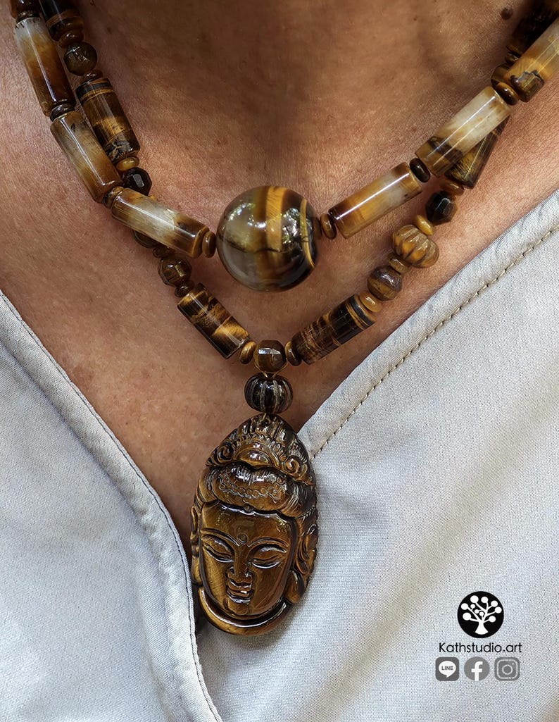Puede incluir: Un collar de doble hebra con cuentas de ojo de tigre pulidas en formas cil&iacute;ndricas y esf&eacute;ricas. La pieza central es un gran colgante de ojo de tigre intrincadamente tallado que representa un rostro sereno. El logotipo Kathstudio.art es visible.