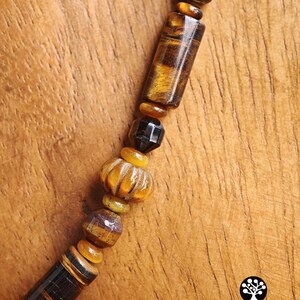 Puede incluir: Un collar con una variedad de cuentas de ojo de tigre en formas cil&iacute;ndricas, facetadas y acanaladas, intercaladas con peque&ntilde;os espaciadores lisos de color &aacute;mbar y una cuenta oscura facetada. El texto Kathstudio.art es visible.