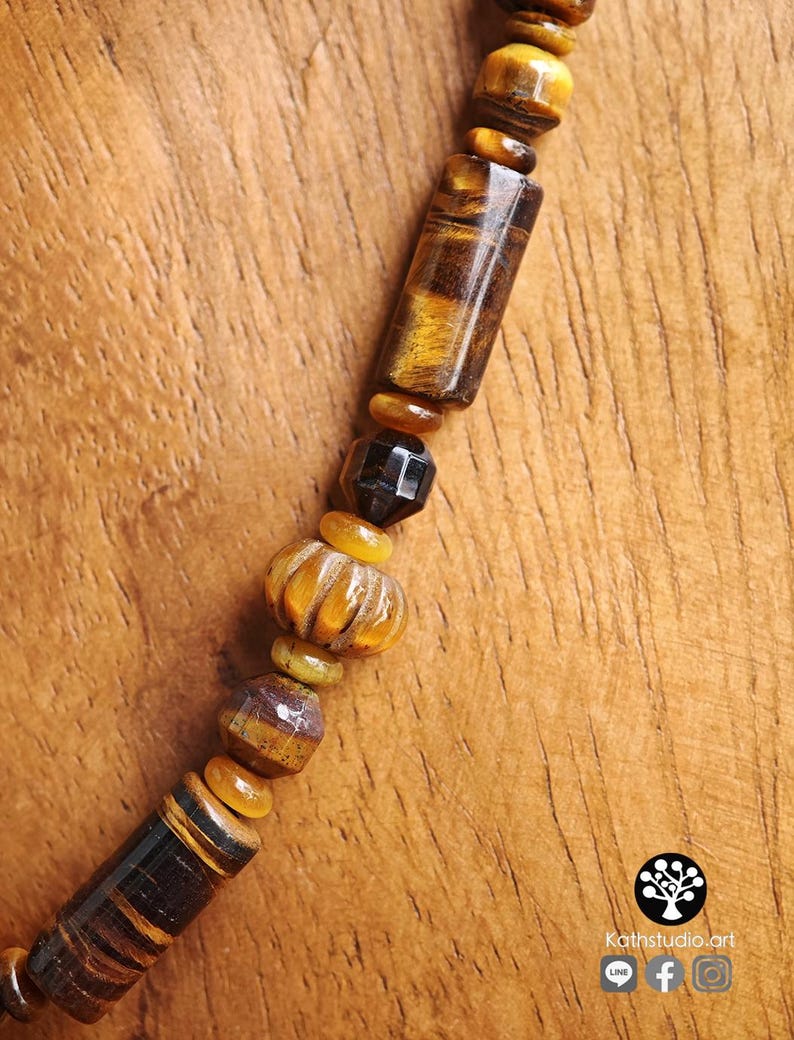 Puede incluir: Un collar con una variedad de cuentas de ojo de tigre en formas cil&iacute;ndricas, facetadas y acanaladas, intercaladas con peque&ntilde;os espaciadores lisos de color &aacute;mbar y una cuenta oscura facetada. El texto Kathstudio.art es visible.