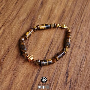May include: A bracelet featuring cylindrical and faceted tiger eye beads in shades of brown and gold. The piece includes a single ornate gold-toned metal spacer bead and a gold-toned metal clasp. The design is arranged in a circular pattern.