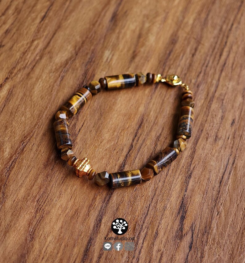 May include: A bracelet featuring cylindrical and faceted tiger eye beads in shades of brown and gold. The piece includes a single ornate gold-toned metal spacer bead and a gold-toned metal clasp. The design is arranged in a circular pattern.