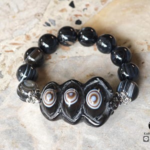May include: A beaded bracelet featuring polished black spherical beads and three central, elongated black beads with white and brown circular eye-like patterns. The central beads are flanked by silver-toned metal filigree accents.