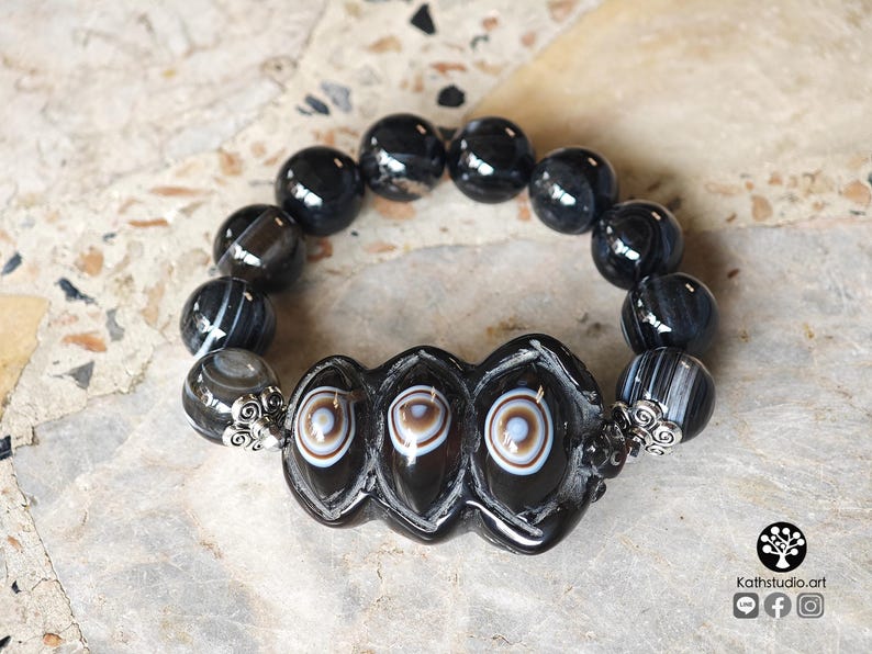 May include: A beaded bracelet featuring polished black spherical beads and three central, elongated black beads with white and brown circular eye-like patterns. The central beads are flanked by silver-toned metal filigree accents.