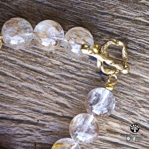 Peut inclure: Un bracelet compos&eacute; de perles sph&eacute;riques en quartz rutile transparent, pr&eacute;sentant des inclusions dor&eacute;es en forme d'aiguilles. Les perles sont reli&eacute;es par un fermoir &agrave; bascule en m&eacute;tal dor&eacute; au design textur&eacute; et torsad&eacute; avec un anneau.