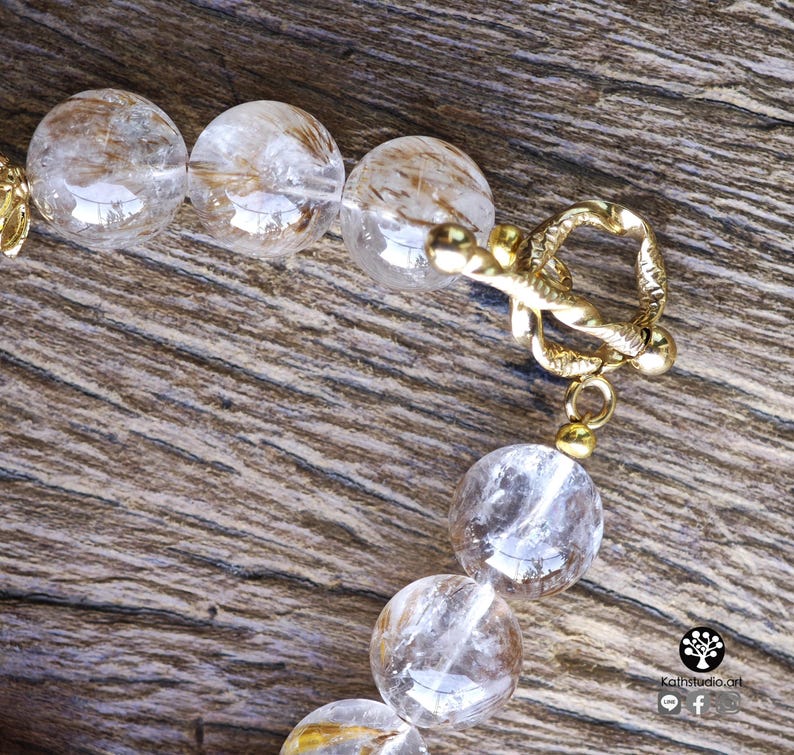 Peut inclure: Un bracelet compos&eacute; de perles sph&eacute;riques en quartz rutile transparent, pr&eacute;sentant des inclusions dor&eacute;es en forme d'aiguilles. Les perles sont reli&eacute;es par un fermoir &agrave; bascule en m&eacute;tal dor&eacute; au design textur&eacute; et torsad&eacute; avec un anneau.