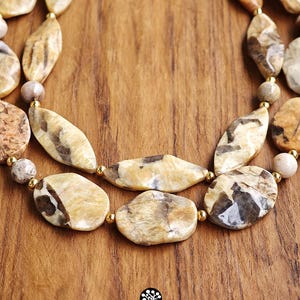 Puede incluir: Un collar de varias hileras con cuentas de piedra pulidas e irregulares en tonos beige y marr&oacute;n, intercaladas con peque&ntilde;as cuentas espaciadoras redondas de color dorado met&aacute;lico. El collar presenta un dise&ntilde;o en capas con formas variadas.