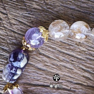 Peut inclure: Un bracelet de perles compos&eacute; de perles d'am&eacute;thyste violette, dont une sculpt&eacute;e en forme de cr&acirc;ne, et de perles de quartz rutile transparent. Les perles sont orn&eacute;es de capuchons en m&eacute;tal dor&eacute;. Un logo avec le texte Kathstudio.art est visible.