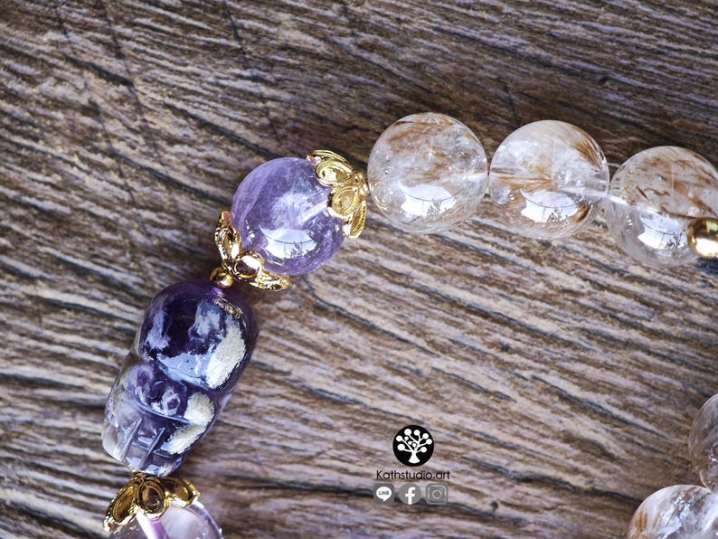 Peut inclure: Un bracelet de perles compos&eacute; de perles d'am&eacute;thyste violette, dont une sculpt&eacute;e en forme de cr&acirc;ne, et de perles de quartz rutile transparent. Les perles sont orn&eacute;es de capuchons en m&eacute;tal dor&eacute;. Un logo avec le texte Kathstudio.art est visible.