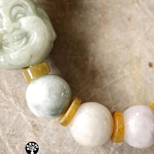 Op de afbeelding: Een armband met een uitgesneden lichtgroene jade Boeddha-kraal, afgewisseld met gladde ronde witte en licht lavendelkleurige jade kralen, gescheiden door kleine platte gele jade tussenstukjes. Het logo KATHSTUDIO BANGKOK NG HERITAGE INTO EVERYDAY ELEGANCE is zichtbaar.