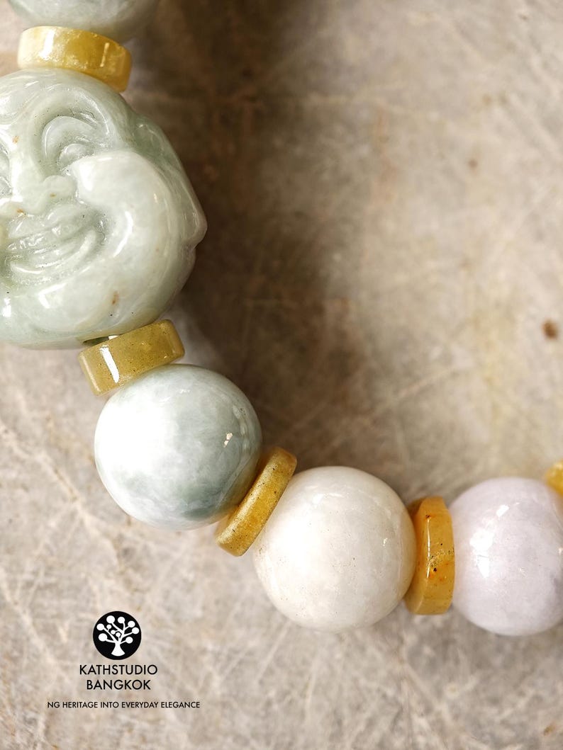 Op de afbeelding: Een armband met een uitgesneden lichtgroene jade Boeddha-kraal, afgewisseld met gladde ronde witte en licht lavendelkleurige jade kralen, gescheiden door kleine platte gele jade tussenstukjes. Het logo KATHSTUDIO BANGKOK NG HERITAGE INTO EVERYDAY ELEGANCE is zichtbaar.