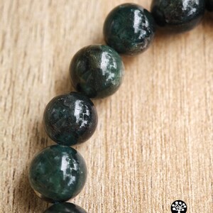 May include: A bracelet composed of smooth, spherical dark green beads with visible internal mineral inclusions. The beads are arranged in a curved line. A small logo with the text Kathstudio.art and social media icons is in the corner.