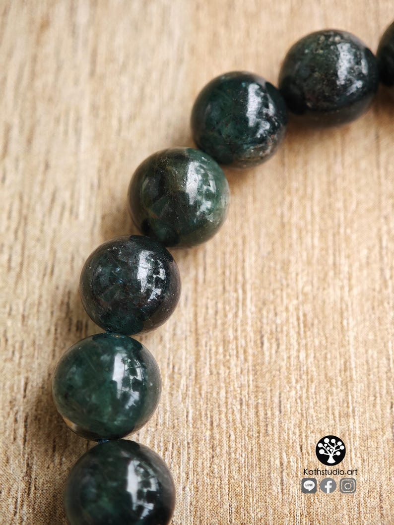 May include: A bracelet composed of smooth, spherical dark green beads with visible internal mineral inclusions. The beads are arranged in a curved line. A small logo with the text Kathstudio.art and social media icons is in the corner.