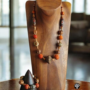 May include: A wooden necklace and bracelet set displayed on a wooden bust and cone. The necklace features various beads in shades of brown, orange, and red. The bracelet has similar beads. The jewelry is displayed on a wooden table.