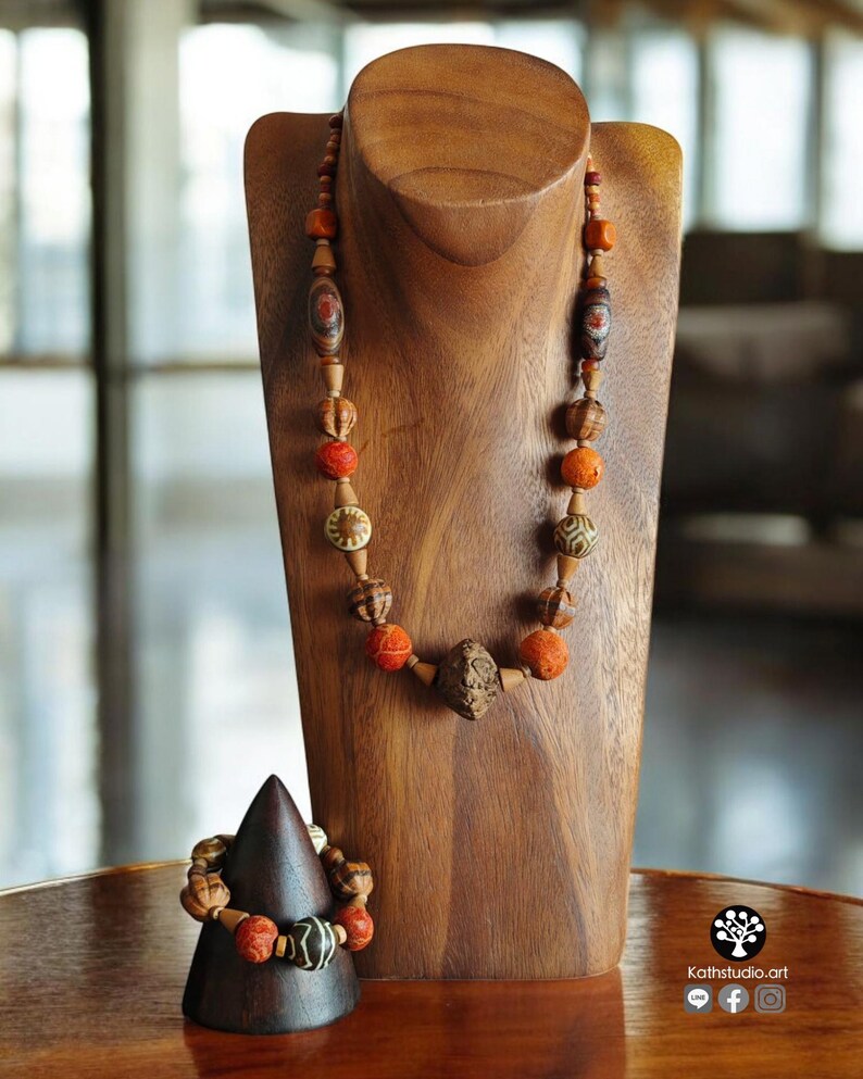 May include: A wooden necklace and bracelet set displayed on a wooden bust and cone. The necklace features various beads in shades of brown, orange, and red. The bracelet has similar beads. The jewelry is displayed on a wooden table.