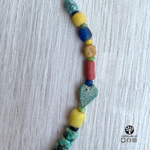 Op de afbeelding: Een kleurrijke ketting met diverse kralen in tinten turquoise, geel, blauw, rood en bruin. De kralen hebben verschillende vormen en maten, waaronder cilindrisch, rond en hartvormig. De ketting wordt getoond tegen een lichte houtnerf achtergrond.