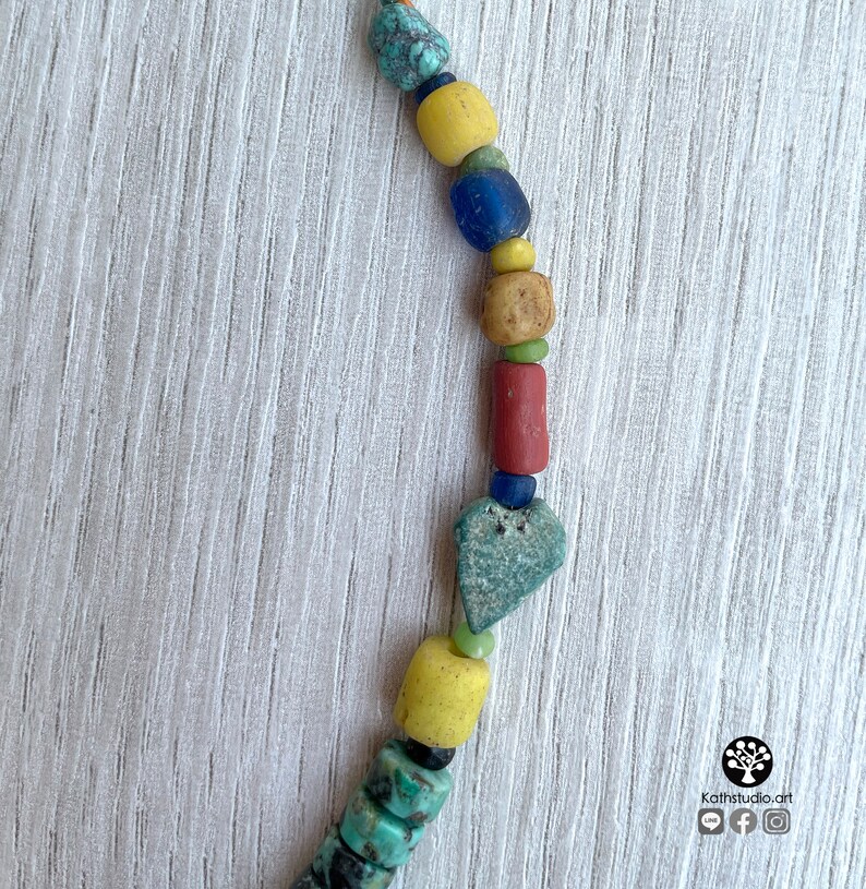 Op de afbeelding: Een kleurrijke ketting met diverse kralen in tinten turquoise, geel, blauw, rood en bruin. De kralen hebben verschillende vormen en maten, waaronder cilindrisch, rond en hartvormig. De ketting wordt getoond tegen een lichte houtnerf achtergrond.