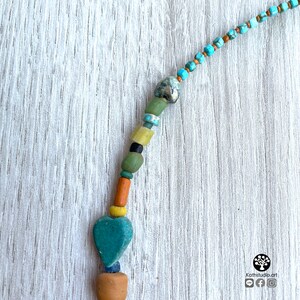 Op de afbeelding: Een handgemaakte ketting met diverse kralen in tinten turquoise, groen, geel, oranje en bruin. De ketting heeft een turquoise hartvormige hanger en een kleine, lichtbruine kleikraal aan de onderkant.