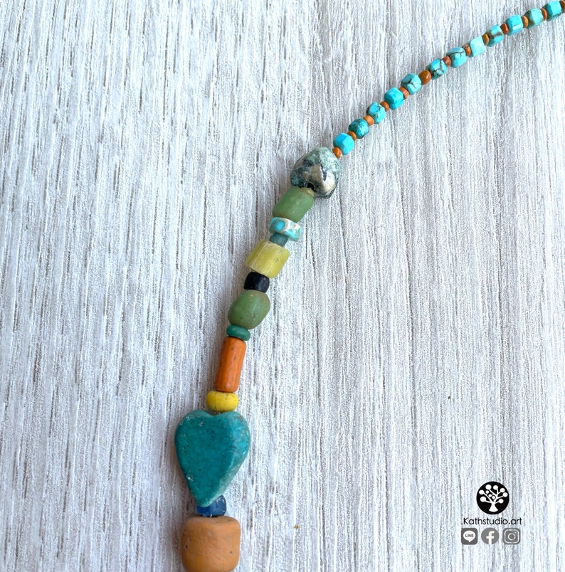 Op de afbeelding: Een handgemaakte ketting met diverse kralen in tinten turquoise, groen, geel, oranje en bruin. De ketting heeft een turquoise hartvormige hanger en een kleine, lichtbruine kleikraal aan de onderkant.