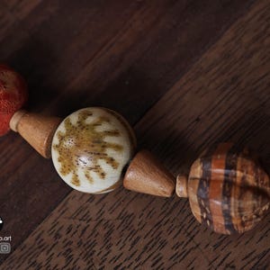 May include: Close-up of a handmade necklace featuring a variety of natural beads. The necklace includes round, patterned beads in shades of brown, cream, and orange. The beads are strung together with wooden connectors, creating a unique and earthy design.