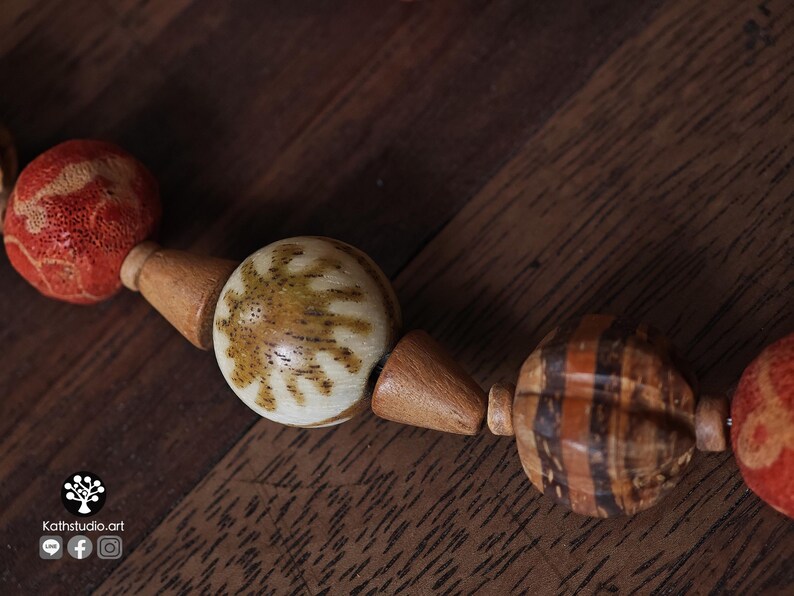 May include: Close-up of a handmade necklace featuring a variety of natural beads. The necklace includes round, patterned beads in shades of brown, cream, and orange. The beads are strung together with wooden connectors, creating a unique and earthy design.