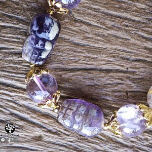 Peut inclure: Un bracelet compos&eacute; de perles d'am&eacute;thyste violettes, dont certaines sont sculpt&eacute;es en forme de cr&acirc;ne, altern&eacute;es avec des coupelles en m&eacute;tal dor&eacute;. Le bijou comporte un petit logo avec le texte Kathstudio.art et des ic&ocirc;nes de r&eacute;seaux sociaux.