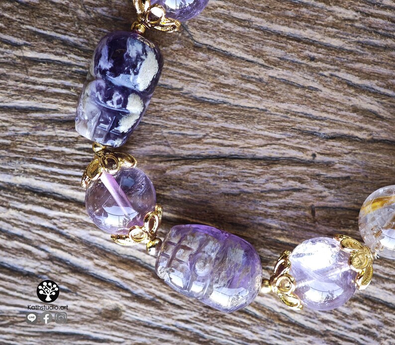 Peut inclure: Un bracelet compos&eacute; de perles d'am&eacute;thyste violettes, dont certaines sont sculpt&eacute;es en forme de cr&acirc;ne, altern&eacute;es avec des coupelles en m&eacute;tal dor&eacute;. Le bijou comporte un petit logo avec le texte Kathstudio.art et des ic&ocirc;nes de r&eacute;seaux sociaux.