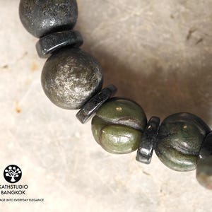 Puede incluir: Una pulsera de cuentas que alterna esferas met&aacute;licas oscuras y cuentas de piedra verde oliva talladas con una textura floral. El logotipo de KATHSTUDIO BANGKOK con el texto TURNING HERITAGE INTO EVERYDAY ELEGANCE es visible en la esquina.