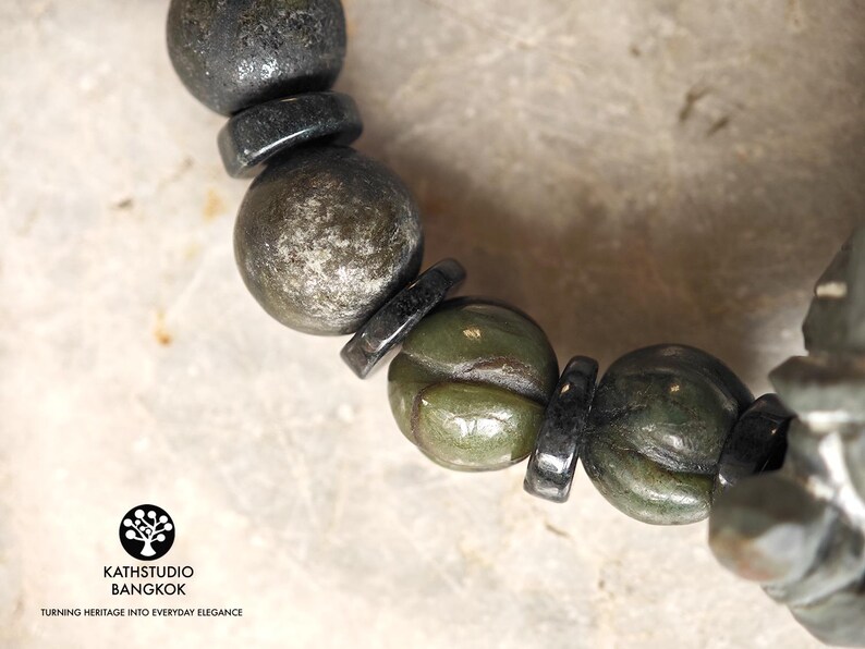 Puede incluir: Una pulsera de cuentas que alterna esferas met&aacute;licas oscuras y cuentas de piedra verde oliva talladas con una textura floral. El logotipo de KATHSTUDIO BANGKOK con el texto TURNING HERITAGE INTO EVERYDAY ELEGANCE es visible en la esquina.