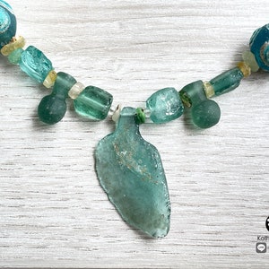 Puede incluir: Collar artesanal con cuentas de vidrio marino en tonos turquesa y transparente. El collar incluye un colgante central con forma de botella. Las cuentas est&aacute;n ensartadas en un cord&oacute;n verde, creando un accesorio &uacute;nico.